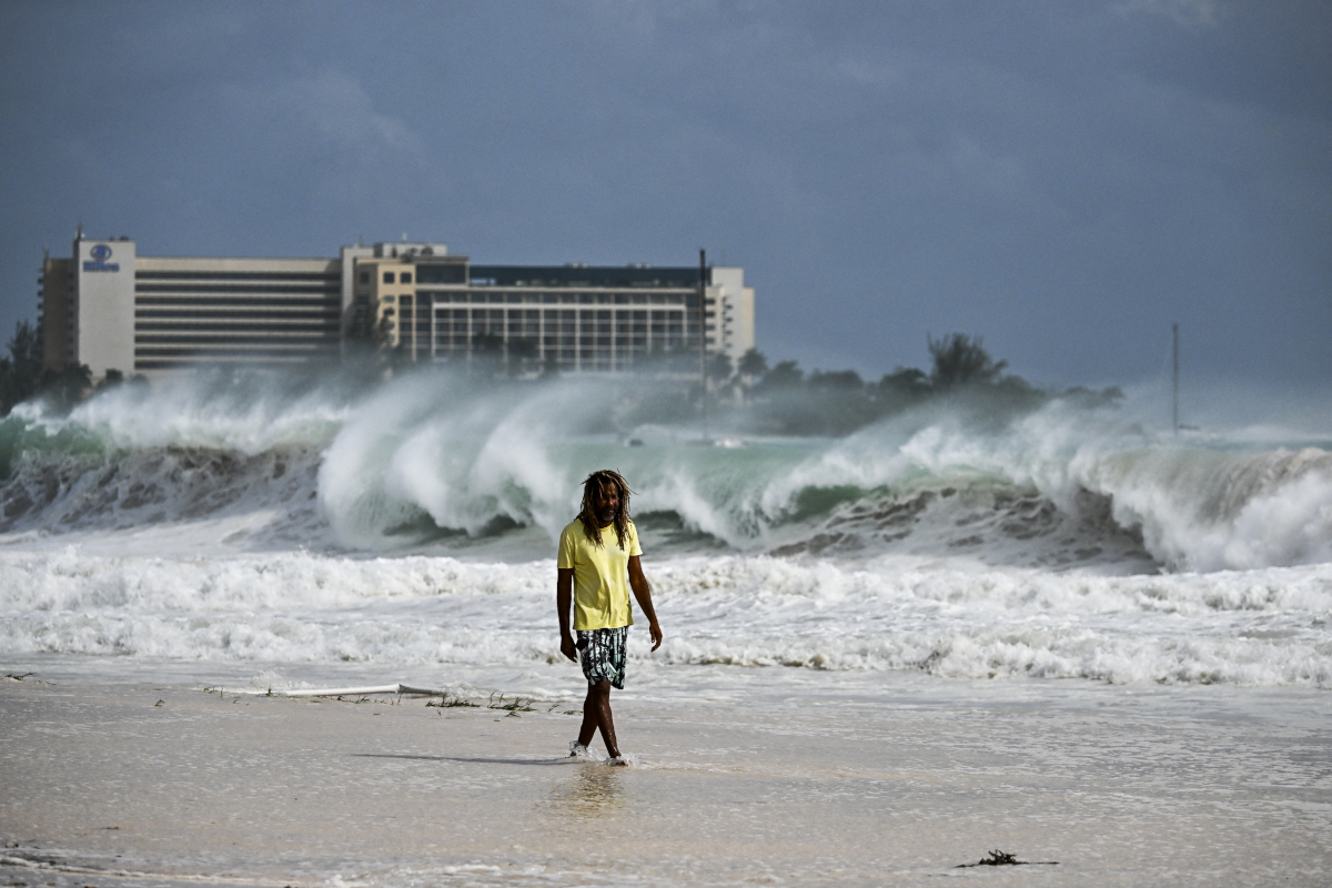 A man walking along a beach with a wave crashing into the sand behind him